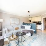 Wohnbereich mit Sofa, Tischen und Küche im Hintergrund.