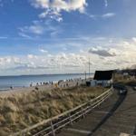 Holzweg entlang der Strandpromenade mit Dünen und Meer im Hintergrund.