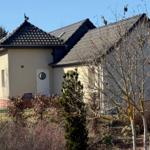 Ein Haus mit cremefarbener Fassade und dunklem Dach, umgeben von Bäumen und Sträuchern.