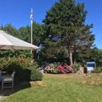 Garten mit Sonnenschirm, Blumenbeete und Liegestühlen unter blauem Himmel.