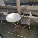 Balkon mit runden Tisch und zwei Stühlen, Hintergrund mit Bäumen und Gebäude.