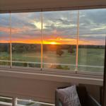 Blick auf Sonnenuntergang über Grünfläche durch großes Fenster mit Sitzgelegenheit.