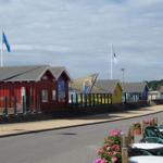 Strandpromenade mit bunten Hütten und Tischen. Blick auf Wasser und Flaggen.
