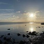 Sonnenlicht reflektiert auf ruhigem Wasser neben einem steinigen Ufer.