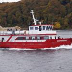 Rotes Schiff mit weißen Aufbauten fährt auf dem Wasser. Menschen stehen auf dem Deck.