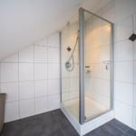 Dusche mit Glaswand, weiße Fliesen, dunkler Boden und Abfalleimer.