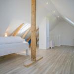 Schlafzimmer mit Dachschrägen, Holzboden und Fenster. Bett, Sitzgelegenheit und Holzstütze sichtbar.