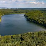 Ein See umgeben von dichtem Wald. Im Hintergrund sind Gebäude sichtbar.