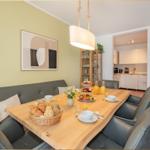 Essbereich mit Holztisch, grauen Stühlen und Frühstücksausstattung. Küche im Hintergrund.