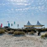 Strand mit Dünen, weißen Zelten und Flaggen unter blauem Himmel.