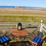 Terrasse mit Tisch und zwei Stühlen mit blauen Kissen. Blick auf Grünfläche und Meer.