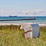 Weißer Strandkorb steht im Gras. Im Hintergrund See, Pier und Schiffe.