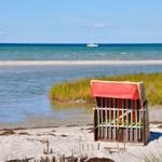 Ein roter Strandkorb steht am sandigen Ufer. Im Hintergrund sind Segelboote auf dem Meer zu sehen.