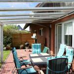 Terrasse mit Sitzgruppe, Grill und Glasdach. Roter Kiesweg und grüner Rasen.