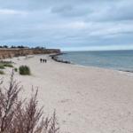 Strand mit Sand, Felsen und wenigen Personen am Wasser.