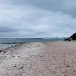 Strand mit Sand, Felsen und Brücke im Hintergrund unter bewölktem Himmel