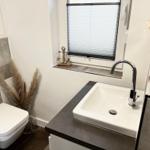 Badezimmer mit Waschbecken, Toilette und Fenster mit Jalousie.