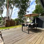 Holzterrasse mit Tisch, Stühlen und Sonnenschutz unter Bäumen.