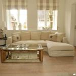 Wohnzimmer mit L-förmigem Sofa, Glas-Tisch und Fenstern mit Rollläden.