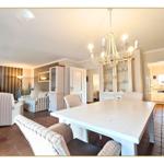 Großes Esszimmer mit weißem Tisch und Stühlen. Wohnbereich mit Sofa und Fernseher. Küche im Hintergrund.