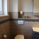 Badezimmer mit Toilette, Waschbecken und Spiegel. Wandfliesen in Braun- und Schwarzton.