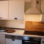 Küche mit Spüle, Kochfeld und Backofen. Holzrückwand und weiße Schränke.