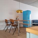 Essbereich mit Tisch, Stühlen und blauem Kühlschrank. Sofa und goldener Hängelampe sind sichtbar.