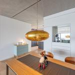 Essbereich mit Holztisch, Stühlen und goldener Hängelampe. Weiße Schrankwand und Küchenbereich im Hintergrund.