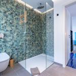 Bad mit Dusche und tropischen Fliesen. Durchgang zu Küche mit blauem Kühlschrank.