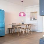 Essbereich mit Holztisch, Stühlen und blauem Kühlschrank unter einer rosa Lampe.