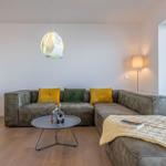 Ein Wohnzimmer mit grauem Sofa, gelben und grünen Kissen, runden Tisch und großen Fenster.