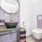 Badezimmer mit Lavendelfliesen, runder Spiegel, schwarzer Waschschale und Wandtoilette.