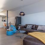 Wohnzimmer mit braunem Sofa, blauem Sessel und Holzboden. Küche und Treppe sind sichtbar.