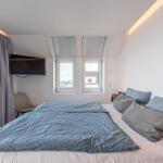 Schlafzimmer mit Bett, Fenstern und TV. Blick auf Wasser durch Glastür.