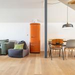 Wohn- und Essbereich mit Sofa, Sitzkissen, orangefarbener Kühlschrank und Holztisch mit Stühlen.