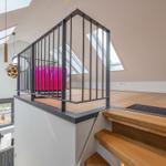 Helle Holztreppe führt zu einer Galerie mit schwarzem Geländer und pinkem Sitzmöbel.