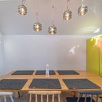 Essbereich mit Holztisch, Stühlen und gelbem Kühlschrank. Deckenleuchten und Platten.