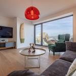 Wohnzimmer mit Sofa, Sessel und TV. Große Fensterfront mit Aussicht. Roter Hängelampe und Holzboden.