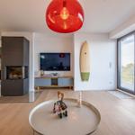 Wohnzimmer mit Kamin, TV, Surfbrett und großem Fenster. Roter Hängelampe und runder Tisch.