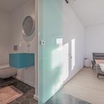 Bad mit Toilette, Waschbecken und Dusche. Nebenraum mit Liege und Fenster.