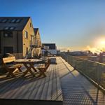 Holzterrasse mit Tisch und Stühlen. Blick auf Gebäude und Sonnenuntergang.