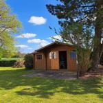 Wooden cabin with open door, surrounded by grass and trees under blue sky.