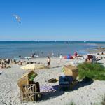 Strand mit Wickerstühlen, Sonnenschirmen und Badegästen am Wasser.