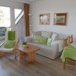 Wohn- und Essbereich mit Sofa, Stuhl und Tisch. Grüne Tischdecke und Kissen. Fenster mit Blick auf Balkon.