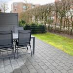 Terrasse mit Tisch und vier Stühlen, gepflastert, mit Rasen und Hecke im Hintergrund.