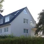 Modernes Haus mit Balkon und Dachfenstern, umgeben von Bäumen und Sträuchern.