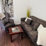 Wohnzimmer mit Sofa, Sessel und Holztisch. Wandbild und Pflanze sind sichtbar.