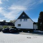 Modern house with white facade and black roof, paved forecourt with parked cars.