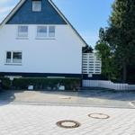 House with white facade and dark roof, paved yard with hedge and tree.