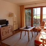 Wohnzimmer mit Sofa, Tisch, Stühlen und TV. Große Glastüren führen auf eine Terrasse.